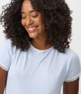 Blusa de pijama de mujer color celeste, confeccionada en tejido peach touch de poliéster con elastano, con estampado de moños sutiles. Presenta cuello redondo, mangas cortas con detalles de encaje blanco y un pequeño moño decorativo en el escote.