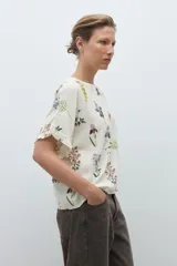 Blusa de manga corta, color crudo, con estampado floral en tonos azules, amarillos y rojos. Presenta cuello redondo y volados en los puños.