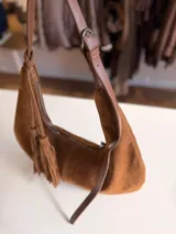 Cartera tipo hobo de textura gamuzada en color marrón oscuro, con diseño flexible y asa larga regulable. Presenta un detalle de flecos largos desmontables en uno de los laterales y cierre superior.