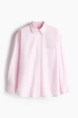 Camisa de corte holgado confeccionada en popelina de algodón, con cuello clásico, cierre frontal de botones, hombros caídos y bolsillo de parche en el pecho. Presenta mangas largas con puños abotonados, canesú en la espalda y bajo redondeado con aberturas laterales, siendo ligeramente más larga en la parte posterior.