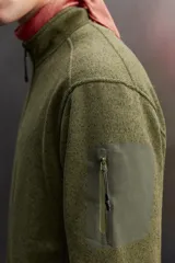 Chaqueta verde militar de punto con interior de polar. Tiene cuello alto, manga larga con bolsillo con cierre en la manga, bolsillos laterales con cierre y cierre frontal con cremallera.