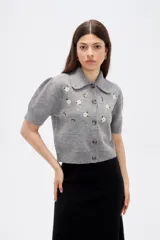 Saco de punto gris con cuello tipo camisa, manga corta y cierre frontal con botones. Presenta un delicado bordado de flores blancas en la parte superior delantera.