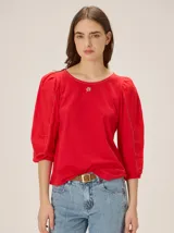 Blusa roja con mangas abullonadas tres cuartos y cuello redondo. Detalle de bordado pequeño en el centro del escote y costuras decorativas visibles en las mangas.