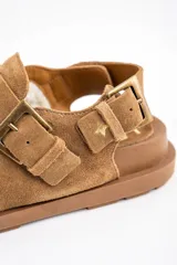 Zueco de gamuza color camel, con interior forrado en corderito, tira ajustable en el talón y suela de goma plana.