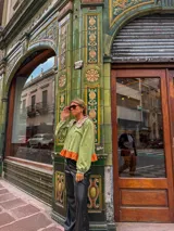 Camisa de manga larga en color verde oliva, con cuello babero en tono contrastante y detalle de volado en el ruedo en color naranja. Presenta cierre frontal con botones y mangas abullonadas con puños ajustados.