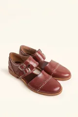 Zapato tipo oxford abierto en los laterales, confeccionado en cuero vacuno negro. Presenta doble tira con hebillas metálicas en el empeine y detalle de tachas plateadas en el contorno del talón y la puntera.