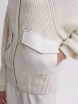 Cárdigan tejido color crema con cierre frontal de cremallera dorada, cuello alto y mangas largas raglán. Presenta dos bolsillos frontales de cuero sintético con solapa y botón a presión.