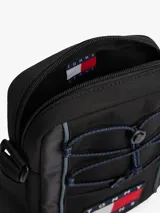 Cartera reporter de estilo urbano color negro, con compartimento principal con cierre, cordones elásticos ajustables en el frente y correa ajustable. Presenta el logo de la marca en un parche frontal y etiqueta lateral de Cordura.