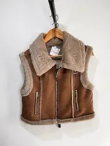 Chaleco estilo gamulán con interior y bordes de piel sintética. Presenta cierre frontal metálico, bolsillos laterales con cierre y detalles de costuras visibles.