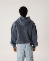 Hoodie de color azul grisáceo con efecto pigmentado/lavado, corte holgado y capucha. Presenta un pequeño bordado frontal con texto y dos estrellas. Posee bolsillo canguro con bordes desgastados.