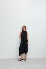Vestido midi de color marrón con acabado satinado, cuello redondo con detalle fruncido y diseño sin mangas. Presenta un corte fluido con bajo asimétrico y bolsillos laterales ocultos.