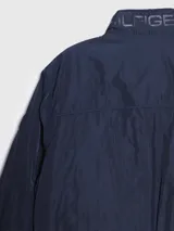 Campera bomber Tommy Hilfiger azul marino con cuello alto, cierre frontal con cremallera, dos bolsillos laterales con cremallera, puños y bajo elásticos. Tiene el logo de Tommy Hilfiger bordado en el pecho y un parche con el logo en la manga izquierda. El interior del cuello tiene detalles en rojo, blanco y azul.