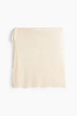 Pañuelo de cadera corto confeccionado en encaje de color beige, con tiras finas para atar en un lateral y terminaciones en ondas.