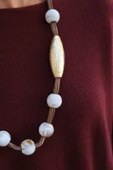 Collar largo de diseño artesanal compuesto por una cuerda central de la que cuelgan cuentas esféricas de cerámica blanca con vetas y dos piezas centrales alargadas con acabado dorado texturizado.