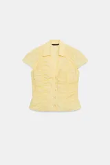 Top de popelín color amarillo, con cuello solapa, escote en pico y manga corta. Presenta un diseño entallado con detalle de frunces verticales en el delantero y cierre frontal con botones.