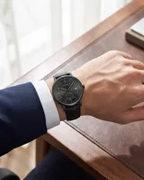 Reloj analógico de pulsera con caja redonda de metal negro, esfera minimalista con indicadores de hora, ventana de fecha y subesfera de segundos. Cuenta con correa de cuero negro y cierre de hebilla.