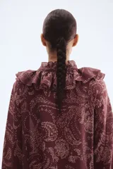 Vestido largo de manga globo, color marrón oscuro, con estampado paisley en tonos claros. Presenta cuello alto con volados y escote en V con lazos ajustables, y puños con volados.