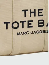 Bolso tipo tote de lona color negro con letras blancas bordadas en el frente que dicen 'THE TOTE BAG MARC JACOBS'. Cuenta con asas superiores cortas y una correa de hombro ajustable y extraíble.