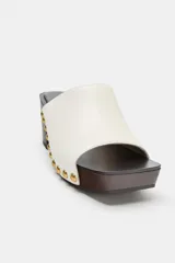 Zueco de piel color blanco, con tacón de madera y plataforma, detalle de tachas metálicas doradas en los laterales y punta cuadrada.