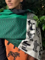 Pashmina de textura suave con diseño de paneles estampados que combinan motivos geométricos en zigzag y figuras de caballos. Presenta una paleta de colores vibrantes que incluye verde, naranja, negro y beige.