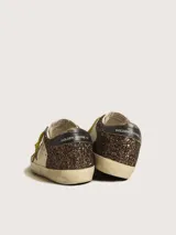 Zapatillas deportivas Golden Goose modelo Super-Star, con cuerpo principal de cuero color crema y detalles de glitter marrón oscuro. Presentan la estrella lateral y el talón en glitter marrón oscuro, cordones amarillos y suela desgastada.