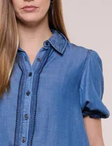 Camisa de lyocell color azul con efecto denim, cuello camisero, cierre frontal con botones y detalles de bordado oscuro a lo largo de la botonera. Presenta mangas cortas abullonadas con puños elásticos y terminación con volados.