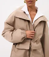 Campera cortaviento oversized color beige, de cuello alto, mangas largas con puños elásticos, bolsillos frontales con solapa y cordón ajustable en la cintura.