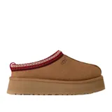 Pantuflas tipo mule UGG modelo Tazz II, confeccionadas en gamuza color tostado con plataforma alta. Presentan un detalle de costura trenzada decorativa en el borde superior en tonos rojo y beige, y el logo de la marca grabado en el lateral.