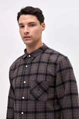 Camisa de corte regular fit, confeccionada en tela de algodón con diseño de cuadros en tonos marrones y negros. Presenta cuello clásico, cierre frontal con botones personalizados y un bolsillo en el pecho con el logo de la marca bordado.