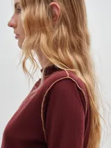 Polera de manga larga confeccionada en tela rib color bordó, con cuello alto y hombreras. Presenta costuras a la vista con pespuntes en hilo de color contrastante en el cuello, puños, hombros y ruedo.