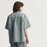 Camisa de jean de manga corta Adidas, confeccionada en 100% algodón, con diseño inspirado en la ropa de trabajo, cierre frontal con broches a presión, cuello camisero, un bolsillo en el pecho con solapa y las clásicas tres tiras de la marca en las mangas.