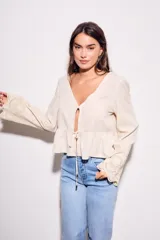 Blusa color beige claro, con escote en V profundo y cierre frontal mediante lazo ajustable a la altura de la cintura. Presenta un peplum con volado y mangas largas con puños anchos con bordados calados.