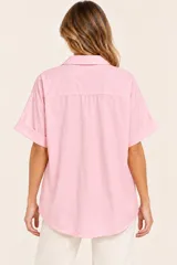 Camisa de algodón color rosa con cuello camisero, escote en V con botón decorativo dorado, mangas cortas con vuelta y diseño holgado.