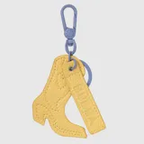 Charm para bolso con forma de bota vaquera y etiqueta rectangular con el logo de la marca grabado, ambos en material sintético texturizado color amarillo. Incluye un mosquetón y una argolla metálica en color lila.
