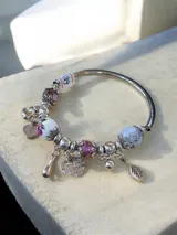Pulsera rígida plateada con dijes colgantes y cuentas decorativas. Incluye cuentas de cristal facetado, cuentas de cerámica con estampado floral, cuentas de piedra veteada y dijes metálicos con forma de corazón, hoja y gota.