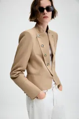 Blazer entallado de sarga color beige con cuello subido y escote en pico. Presenta manga larga con hombreras, detalle de botones metálicos decorativos en el frente y cierre frontal con gancho metálico. Incluye falsos bolsillos de vivo y abertura en la espalda.