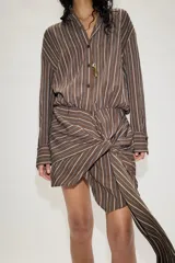 Vestido corto de estilo camisero con estampado de rayas verticales, cuello solapa, manga larga y cierre frontal con botones. Presenta un detalle de lazada en la cintura que crea un efecto drapeado y un bajo asimétrico.