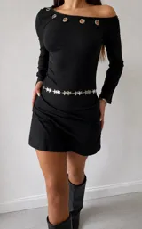 Vestido corto negro de manga larga, con diseño de escote asimétrico decorado con ojales metálicos en la parte superior. Presenta un corte ajustado y una cadena decorativa con dijes de estrellas en la cintura.