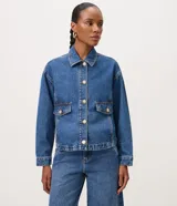 Campera de jean azul de corte recto y holgado, con cuello camisero, cierre frontal de botones metálicos dorados, bolsillos frontales con solapa y mangas largas con puños abotonados.