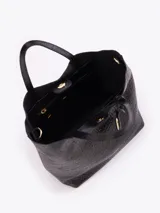 Tote bag Jazmin Chebar de cuero vacuno negro con textura grabada. Presenta un diseño compacto con doble asa superior y un tag colgante con un aplique metálico en forma de labios.