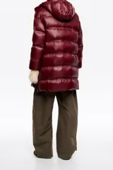 Campera tipo plumífero de largo medio con silueta en línea A, confeccionada en tejido técnico brillante. Presenta cierre frontal con cremallera, cuello subido con capucha desmontable, mangas largas con puños interiores de pelo sintético y bolsillos laterales con cremallera.