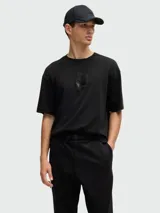 Camiseta negra de corte relajado con cuello redondo y manga corta. Presenta un diseño tridimensional en el pecho que simula un pliegue o estructura geométrica en tono negro brillante o texturizado.