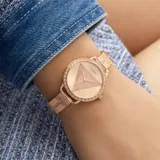 Reloj analógico Guess con caja redonda de 30mm y malla de acero inoxidable en tono oro rosa. La esfera presenta un diseño triangular central con textura brillante y el bisel está decorado con cristales incrustados, al igual que los eslabones iniciales de la correa.
