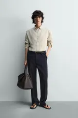 Camisa de algodón de corte relajado, con cuello solapa abotonado (button-down) y manga larga terminada en puño con botón. Presenta un patrón de rayas verticales finas en tonos beige claro y marrón/verde oliva.