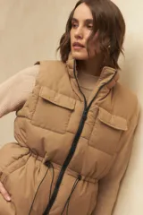 Chaleco puffer color crema, con cuello alto, cierre frontal y cuatro bolsillos con solapa. Cuenta con cintura ajustable con cordón.