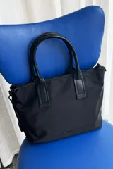 Cartera tipo shopper color negro, de diseño estructurado con doble asa de mano y cierre superior. Presenta sutil grabado de la marca en la parte inferior frontal.