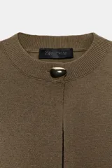 Chaqueta corta color verde oliva, de tejido de punto con cuello redondo y manga corta. Cierre frontal con un único botón dorado.