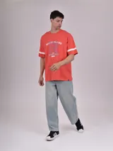 Remera de manga corta color rojo coral, con cuello redondo y detalles de franjas blancas en las mangas. Presenta un estampado frontal con texto que incluye "RHODE ISLAND", "1990" y "VARSITY LEAGUE".