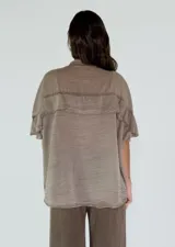 Camisa de textura satinada en tono beige, con cuello mao, cierre frontal de botones y detalle de volado horizontal en el pecho y mangas cortas.