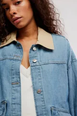 Campera de denim oversize color celeste, con hombro caído, cuello de lona beige en contraste, bolsillos frontales asimétricos, cierre frontal con botones y puños abotonados.